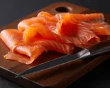 Finas lonchas de salmón ahumado, listas para disfrutar, reposan sobre una tabla de madera junto a un cuchillo afilado.
