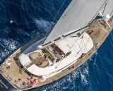Le yacht "Bayesian" navigue sur des eaux bleues, ses voiles blanches gonflées par le vent. L'équipage en rouge s'affaire sur le pont en bois.