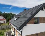 Nowoczesny dom z antracytową elewacją i dachówką, otoczony zielenią i błękitnym niebem.