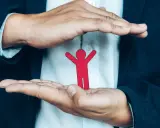 Dłonie obejmujące czerwoną postać symbolizują opiekę i odpowiedzialność za drugiego człowieka.