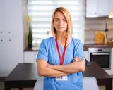 Pielęgniarka środowiskowa NFZ w niebieskim uniformie stoi z rękami skrzyżowanymi na piersi.