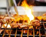 Marinierte Lammkoteletts brutzeln auf dem Grill. Flammen züngeln über dem Fleisch, das perfekt gegrillt wird.