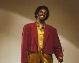 Un percussionniste célèbre, souriant, porte une veste courte bordeaux et une chemise dorée, tenant une baguette.