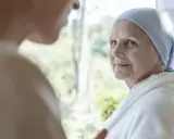 Starsza kobieta w chuście i szlafroku patrzy na kogoś, w tle okno.