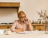 Kobieta w okularach rozmawia przez telefon, siedząc przy stole w kuchni. Przed nią rachunki. Prowadzi **osobne gospodarstwo domowe z mężem**, organizując finanse.