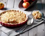 Un délicieux crumble pomme individuel, doré et croustillant, prêt à être dégusté. Des pommes fraîches sont visibles en arrière-plan.