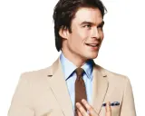 A man in a tan suit and blue shirt smiles, adjusting his tie.