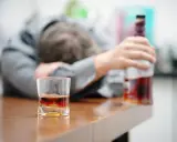 Zaniedbany mężczyzna, prawdopodobnie po eksmisji alkoholika z domu, leży na stole obok szklanki z whisky i butelki.