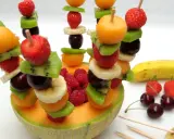 Une **brochette de fruits** colorée avec fraises, kiwis, bananes, cerises et melon, présentée dans une moitié de melon.