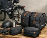 Rowerzysta przygotowuje się do wyprawy, wybierając jakie sakwy rowerowe zabrać. Na stole leżą czarne torby, kask i bidon.