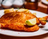 Chrupiący panierowany filet rybny z frytkami i ćwiartką cytryny na białym talerzu.