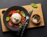 Pyszna burrata z pomidorami, polana oliwą, udekorowana bazylią. Obok miseczka z solą i pieprzem.