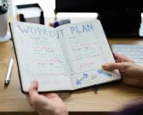 Ein offenes Notizbuch mit einem Trainingsplan, der Übungen und Ruhetage auflistet. Der Plan ist in Spalten für die Tage der Woche unterteilt, mit Kästchen zum Abhaken. Der 3er Split ist klar erkennbar.