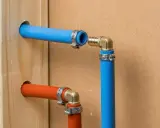 Instalacja hydrauliczna z rurami PEX. Widoczne zaciskanie PEX na złączkach, niebieska i pomarańczowa rura wychodząca z drewnianej belki.