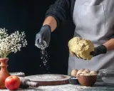 Hände in schwarzen Handschuhen streuen selbsttreibendes Mehl über Teig. Ein Apfel und Blumen sind auf dem Tisch.
