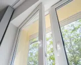 Białe okna z PVC uchylone, wpuszczają do wnętrza światło i zieleń drzew.