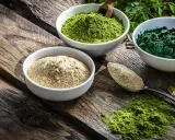 Drei Schalen mit Pulver auf Holz: hellbraun, leuchtend grün und dunkelgrün. Spirulina gesund und voller Nährstoffe.