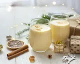 Zwei Gläser cremiger Cocktail mit Eigelb, garniert mit Zimt, umgeben von Weihnachtsdekorationen und Geschenken.
