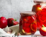 Zwei Gläser mit eingelegter Paprika, daneben frische rote Paprika, Knoblauch und Pfefferkörner. Perfekt zum Paprika einkochen.