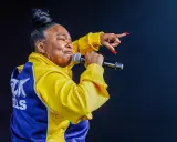 Roxanne Shanté w żółtej kurtce z niebieskimi wstawkami śpiewa do mikrofonu, wskazując palcem.