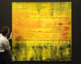 A man in white gloves carefully handles a large abstract painting with vibrant yellow and orange hues, a testament to Gerhard Richter's net worth.