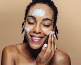 Une femme applique de la crème sur son visage. Elle sourit, les yeux fermés. Elle se demande que veut dire indice 50 pour sa protection solaire.