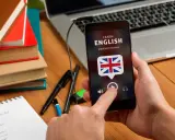 Ręka trzyma smartfon z aplikacją do nauki angielskiego. Na biurku leżą książki i notatnik z nowymi słówkami.