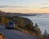 En husbil kör längs en slingrande väg vid den vackra Höga kusten. Solnedgången färgar himlen i mjuka toner över det glittrande havet.