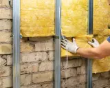 Pose d'isolant jaune sur une structure métallique fixée à un mur de briques. L'isolation intérieure est en cours.