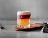 Ein New York Sour mit schichtweisem Rotwein, Whiskey und Schaum. Daneben ein Jigger und ein Barlöffel.