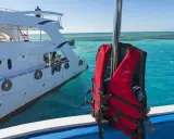 Gilet de sauvetage rouge prêt pour l'aventure en mer, à côté d'un yacht blanc. Les **œuvres mortes du bateau** se reflètent dans l'eau turquoise.