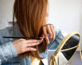 Frau mit langen, braunen Haaren, die sich im Spiegel die Haare schneidet.