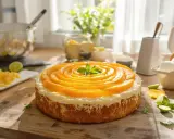 Gâteau sans farine de blé, décoré de rondelles de pêche disposées en spirale et d'une touche de menthe. Des ingrédients comme des œufs et des citrons verts sont visibles sur la table.