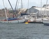 Un voilier blanc et bois, bâché, est partiellement immergé dans un port. Un homme en gilet rouge est à bord.