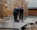 Två personer installerar solceller på ett papptak. Höstfärgade träd syns i bakgrunden.