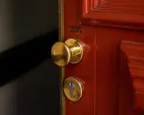 Poignée et serrure dorées sur une porte rouge. Une étiquette indique un service de serrurerie, utile en cas de porte forcée, pour l'assurance propriétaire ou locataire.