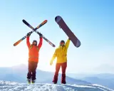 Dwóch narciarzy na szczycie góry, jeden z nartami, drugi z deską snowboardową, cieszy się słońcem i widokiem.