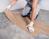 Manos enguantadas colocando suelo vinílico imitación madera sobre suelo de cemento.
