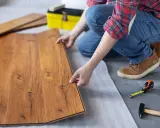 Persona instala tarima flotante, uniendo tablas de madera con cuidado. Herramientas como martillo y cinta métrica a mano.