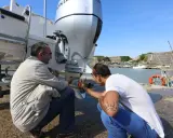 Deux hommes vérifient le montage moteur hors bord sur tableau arrière d'un bateau, près d'un port.