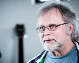 Clark Olofsson idag, med grått hår, skägg och glasögon, blickar mot betraktaren.
