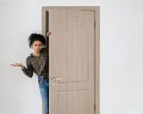 Mujer asomándose por una puerta de madera clara, con expresión de duda. Los colores de puertas como esta son muy versátiles.