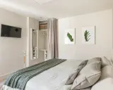 Ein offener Kleiderschrank, der wie ein Kleiderschrank selber bauen mit Vorhang wirkt, mit Regalen und Kleiderstange. Daneben ein Spiegel und ein Bett mit grüner Decke.