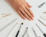 Dłoń z naturalnymi paznokciami otoczona akcesoriami do manicure. Wśród nich pilnik do paznokci naturalnych, cążki, nożyczki i pędzelki.