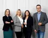 Cztery osoby w strojach biznesowych stoją obok siebie na jasnym tle.