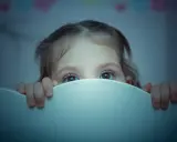 Occhi spalancati nel buio, un bambino si nasconde dietro il bordo del lettino, vittima di un pavor notturno.