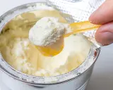 Eine Hand schöpft mit einem gelben Löffel feines Pulver aus einer Dose. Manche denken, dass Proteinpulver dick machen kann, doch es kommt auf die Menge an.
