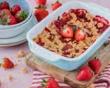 Ein frisch gebackener Erdbeer Crumble in einer hellblauen Auflaufform, garniert mit Erdbeerhälften. Frische Erdbeeren liegen daneben.