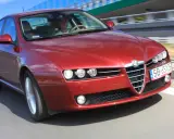 Czerwone Alfa Romeo 159 1.9 JTD pędzi po drodze, kierowca skupiony na trasie.