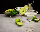 Zwei Margaritas mit Salzrand, garniert mit Limettenscheiben und Minze. Limetten und Minze in einer Schale im Hintergrund. Perfekt für ein Margarita Rezept.